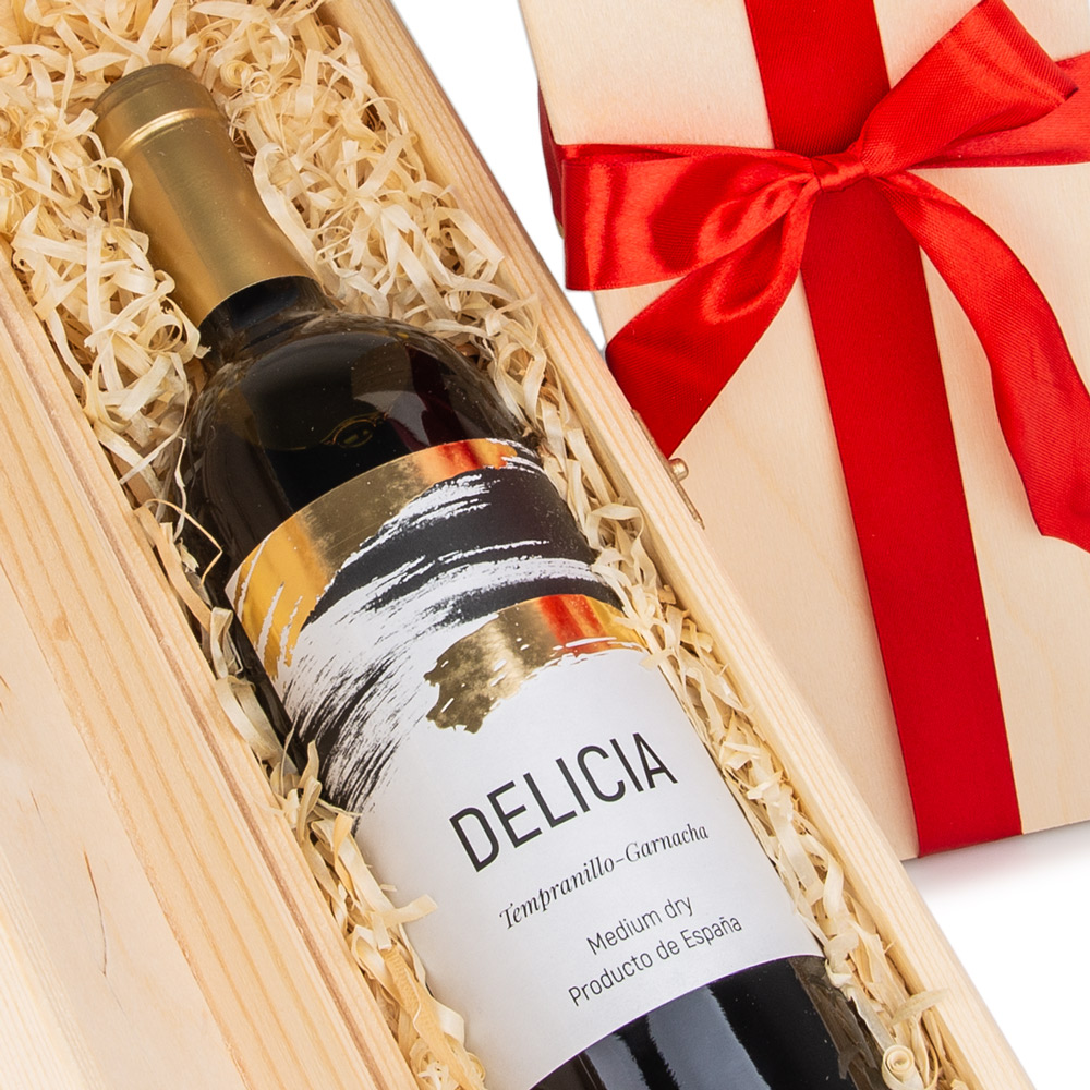 Zbliżenie na butelkę czerwonego wina Delicia Tempranillo-Garnacha leżącą na jasnych wiórkach drewnianych w otwartej, naturalnej drewnianej skrzynce. W tle widoczna górna część zasuniętej skrzynki przewiązanej czerwoną kokardą