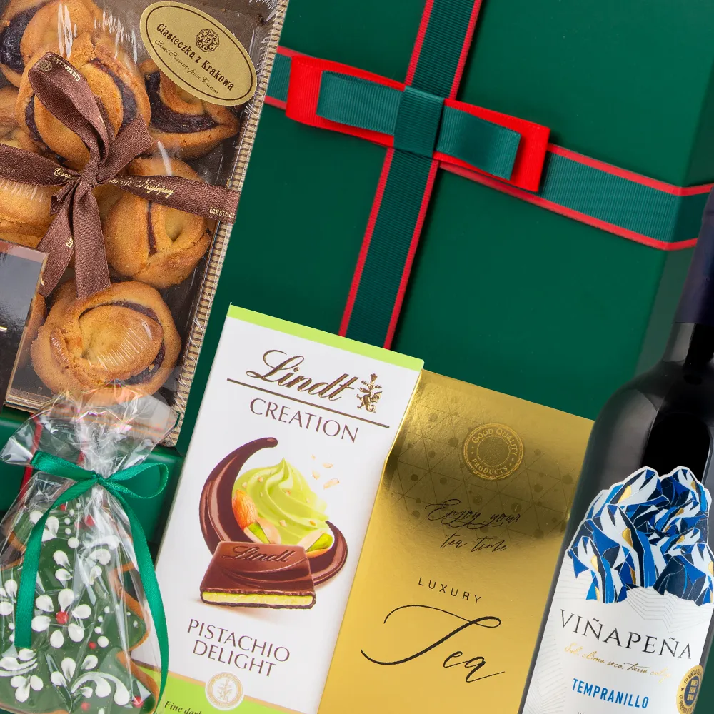 Kompozycja zestawu prezentowego z winem: butelka Vinapena Tempranillo, ciasteczka z różą i Czekolada Lindt Pistachio Delight. Widoczne zielone ozdobne pudełko.